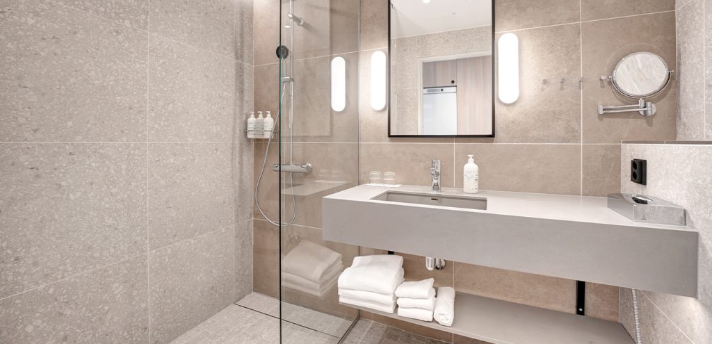 Hotellihuoneen kylpyhuone, jossa on iso kivikuvioitu laatta seinässä, suihkunurkkaus lasiseinän takana ja allaspöydän alla viikattuja pyyhkeitä.