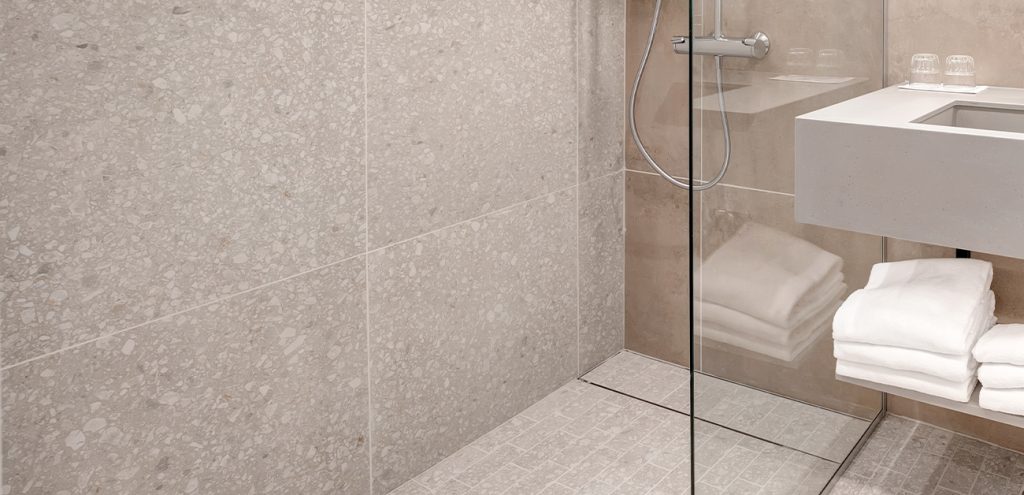 Hotellihuoneen kylpyhuone, jossa on iso kivikuvioitu laatta seinässä, suihkunurkkaus lasiseinän takana ja allaspöydän alla viikattuja pyyhkeitä.